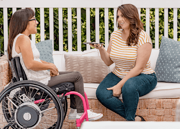 Em um quintal, uma mulher hispânica está sentada em uma cadeira de rodas manual e conversa com a amiga, uma mulher branca segurando um smartphone Android.