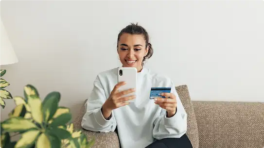 A young woman in a light blue top uses a credit card to complete a payment on her smartphone.