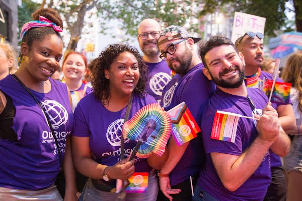 A diverse group of joyful people at an outdoor event wearing purple Outright Action t-shirts, waving Gay Pride rainbow flags