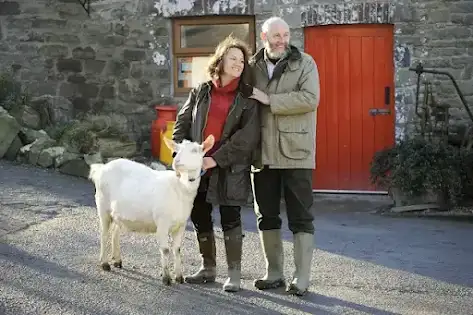 An older man and woman stand next to each other while holding a goat in front of a stone farm building with a red door.