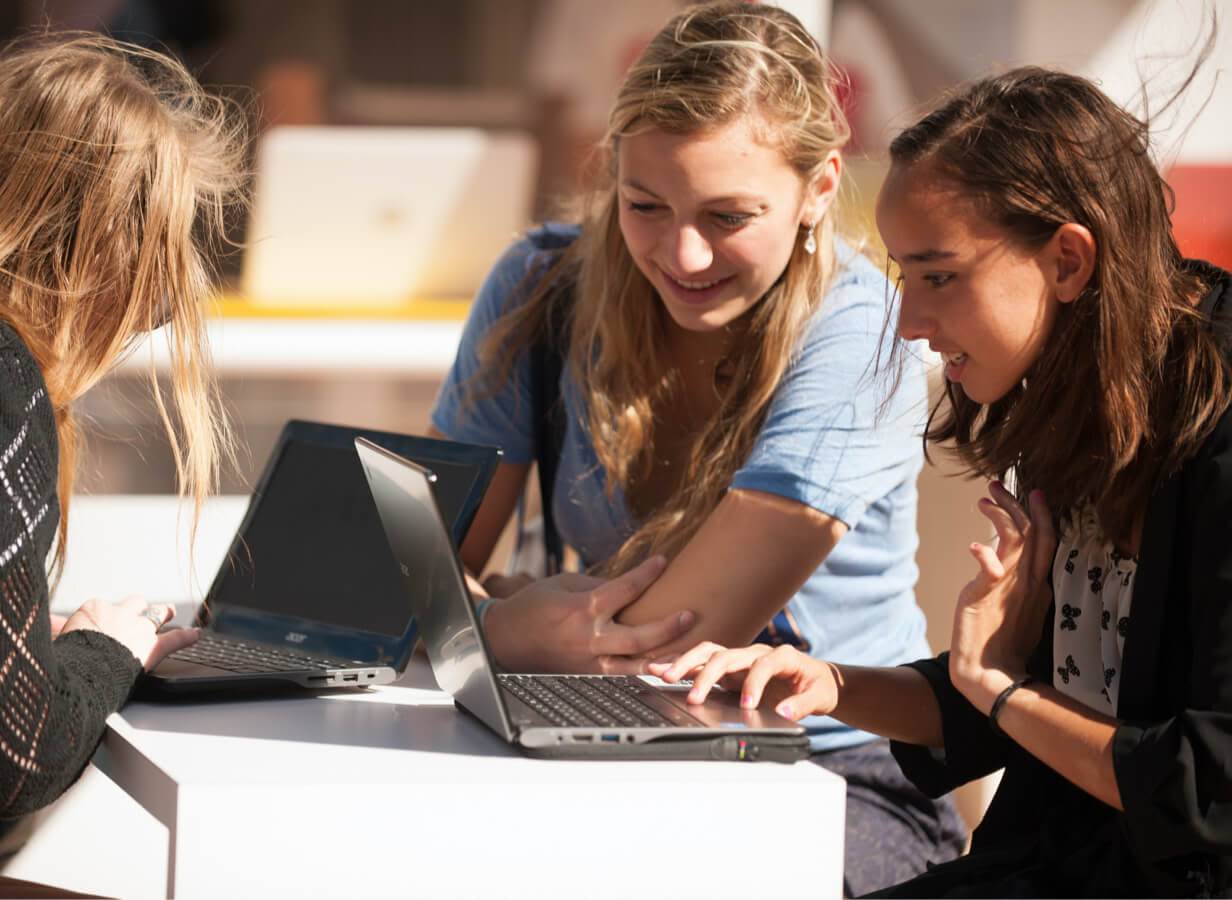 Tiga orang pelajar duduk di meja di luar, membuat kerja menggunakan Chromebook mereka.