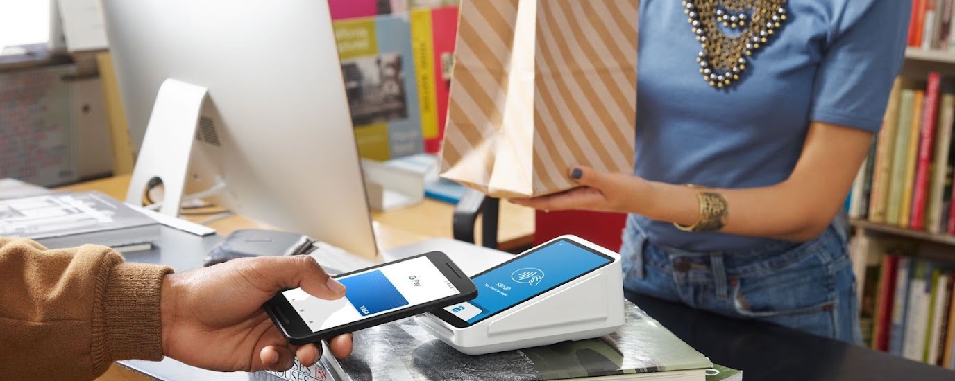 Shopper purchasing with touchless mobile checkout, woman cashier in blue t-shirt hands over diagonally striped paper shopping bag.
