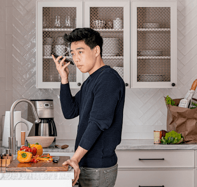 Um homem asiático-americano está no balcão da cozinha enquanto segura um smartphone Android e ouve o leitor de tela verbalizar uma receita.