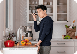 Um homem asiático-americano está no balcão da cozinha enquanto segura um smartphone Android e ouve o leitor de tela verbalizar uma receita.