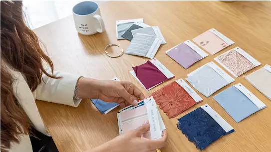 Ali sorts through fabric swatches of different types and colors on a wooden desk.