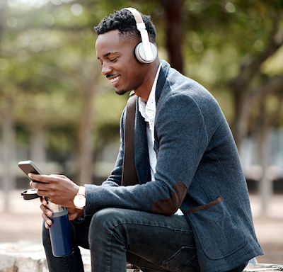 A professional, Black man wearing over-ear headphones is smiling while holding his mobile device and a water bottle while sitting outside on a park bench.