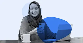 Hidžabą dėvinti moteris šypsosi naudodamasi išmaniuoju telefonu ir skreitiniu kompiuteriu.