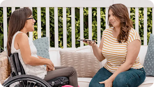 A Hispanic woman sits in her manual push wheelchair and chats with her friend, a white woman holding an Android phone, in a backyard patio.