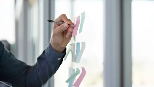 A person’s left arm writes on sticky notes that are placed on a glass wall.