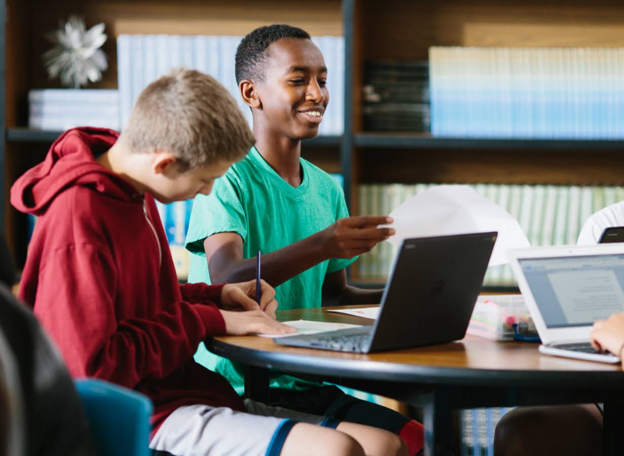 En elev håller i ett papper medan en annan elev i närheten arbetar på en Chromebook.
