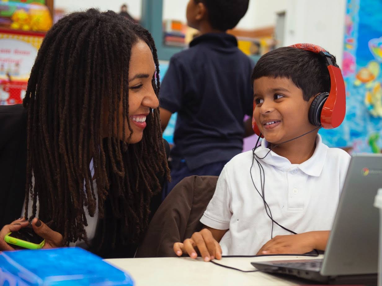 Kobieta nachyla się i uśmiecha do ucznia, który słucha czegoś przez słuchawki podczas korzystania z Chromebooka.