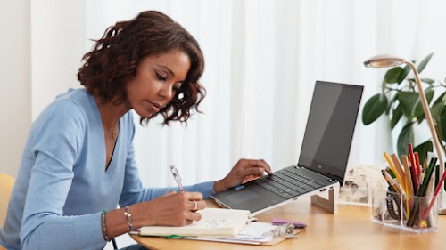 Una mujer sentada frente a un ordenador mientras toma notas en un cuaderno.