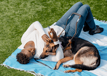 Uma mulher negra deitada em uma toalha de piquenique na grama com o cão de serviço dela, um pastor-alemão, enquanto segura um dispositivo Android na mão.