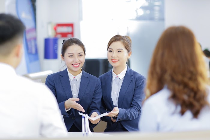 Two young vietnamese women in blue blazers and white blouses behind counter in bank offer two people a brochure