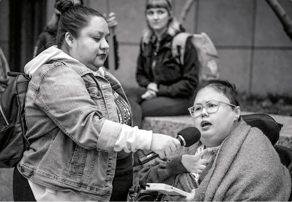 A woman in a wheelchair speaks into a microphone held by a woman standing by her side.