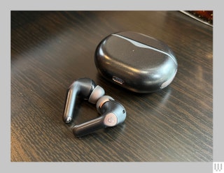 Closed black rounded rectangular case beside two black and grey earbuds all sitting on a dark surface