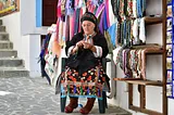 An Eastern-European woman in colorful garb sits outside a store and sews