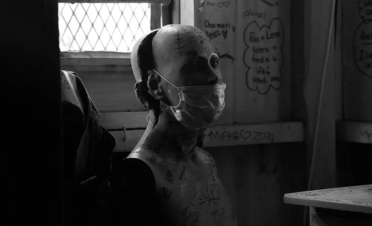 A mannequin that has been partially dismembered, sitting near a window and against a wall with graffiti. It has a surgical mask on its face.