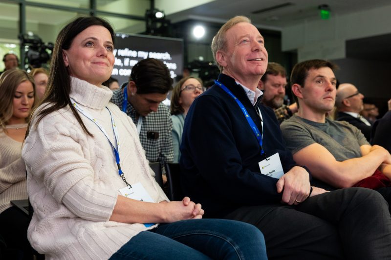 Microsoft executives smile as they watch a news briefing unfold onstage.