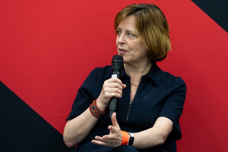 Melanie Dawes speaking into a microphone in front of a red and black background.