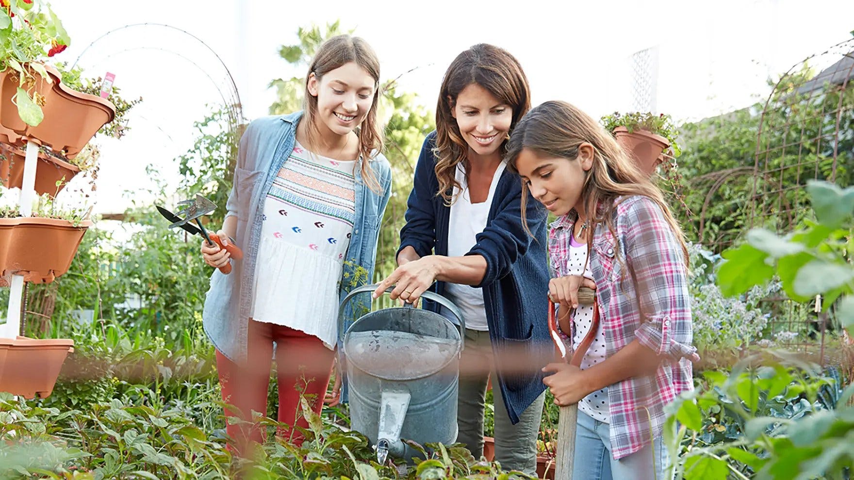 A mother and two preteen girls holding gardening tools surrounded by plants