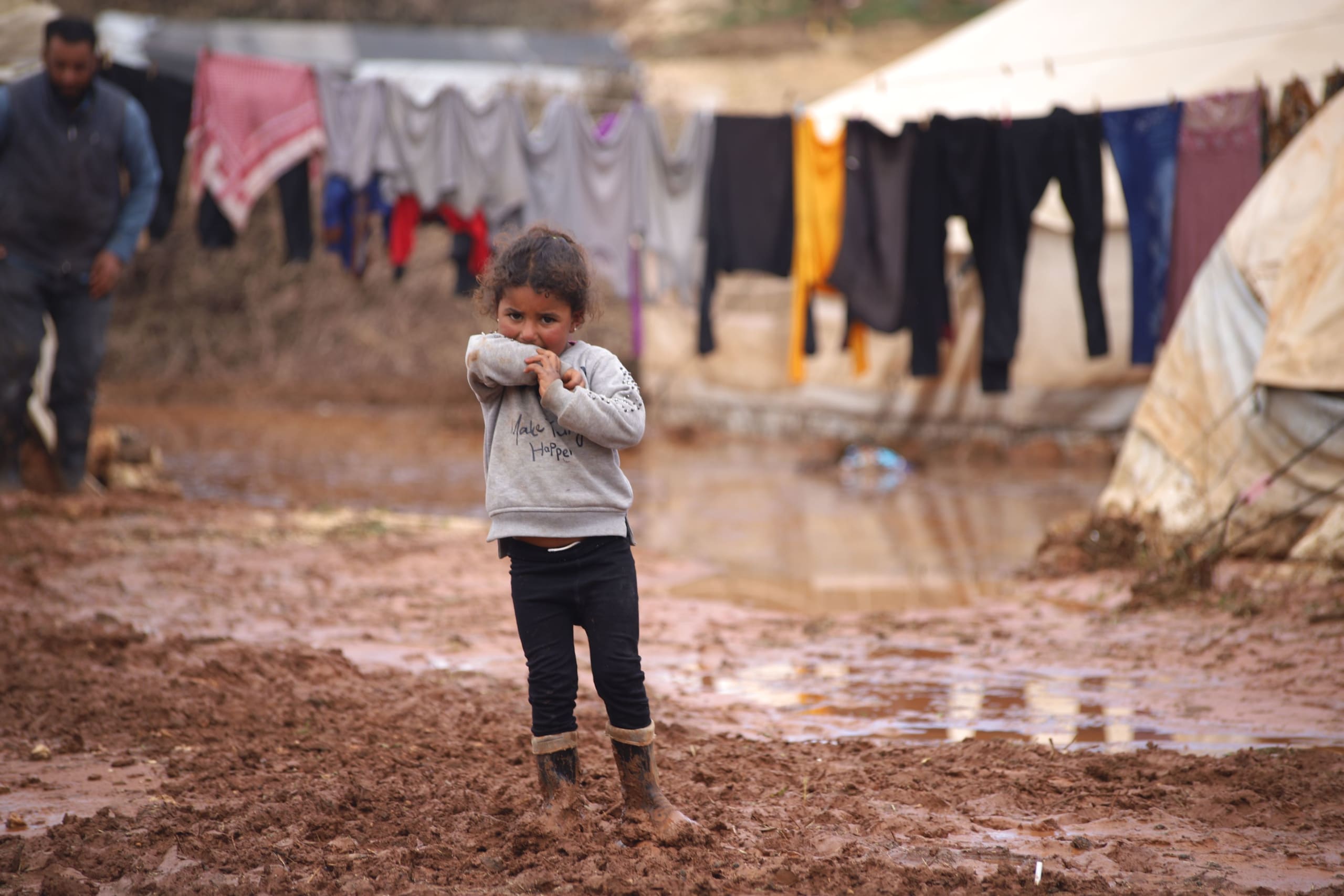 En lille syrisk pige står foran sin families telt i en flygtningelejer for internt fordrevne. Pigen er omringet af vand og står i mudder til anklerne, som følger af oversvømmelser i lejren.