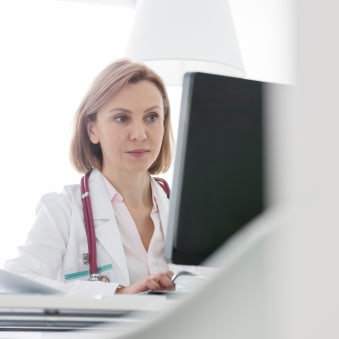 A doctor in a white coat with a stethoscope sits at a desktop computer.