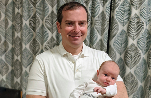 ="Selig holds his infant child, who is wearing a Meta onsie"