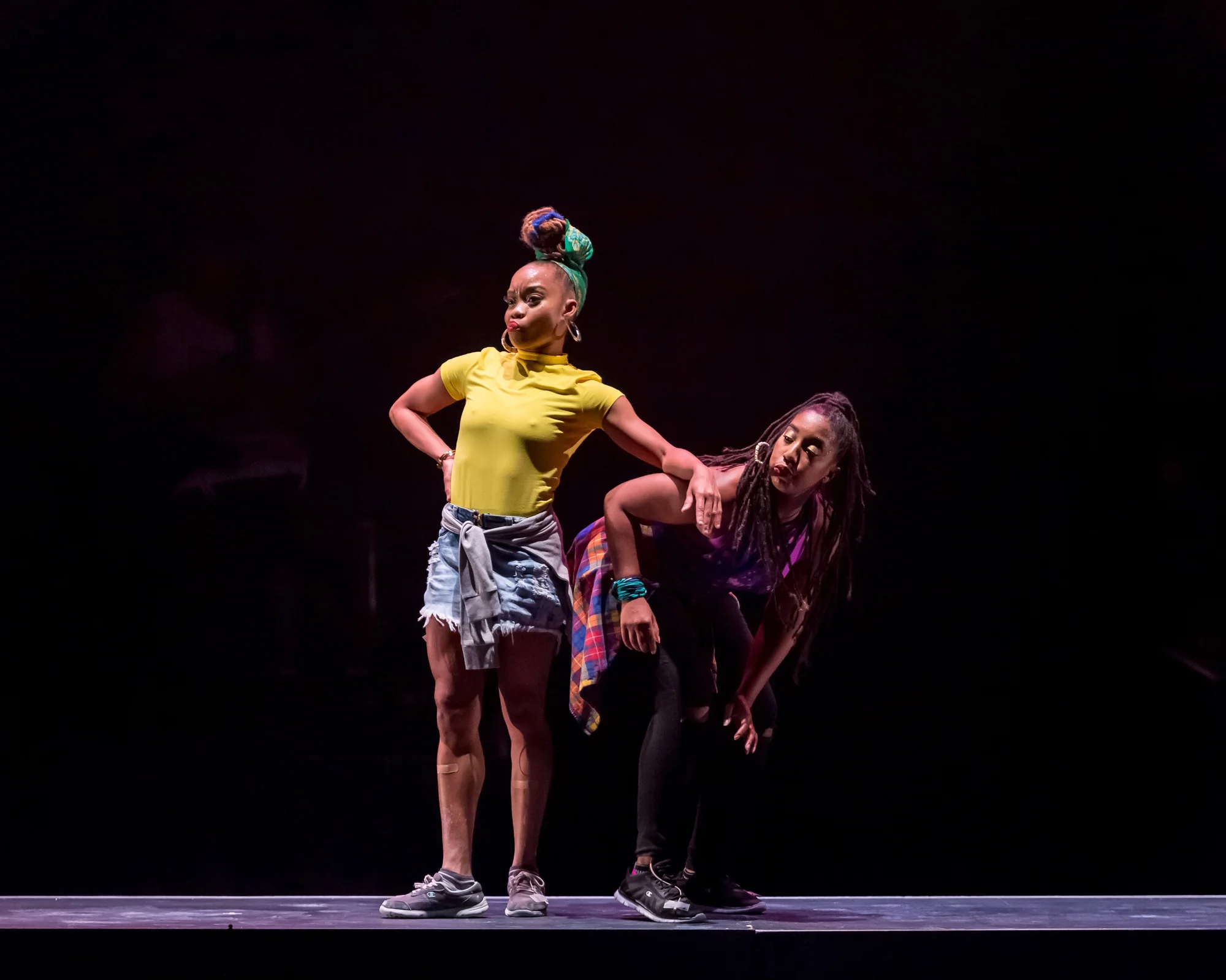 Color photograph of two women on a stage. One woman is bent over, seemingly in a dance position. She wears black leggings, a purple top, and a plaid top tied around her waist. The second woman keeps her arm on the other woman’s shoulder. She wears a yellow shirt, denim shorts, and sneakers.