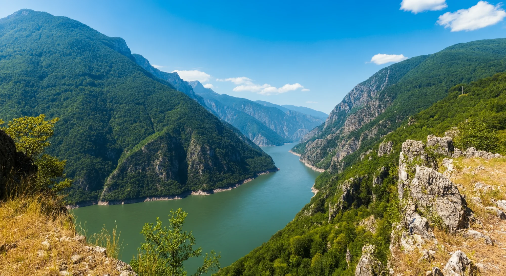 A wide river winds through a deep gorge carved into a lush, green mountain range under a clear blue sky. The river is calm and reflects the surrounding landscape. The sun shines brightly, casting shadows on the slopes and highlighting the textures of the rocks.