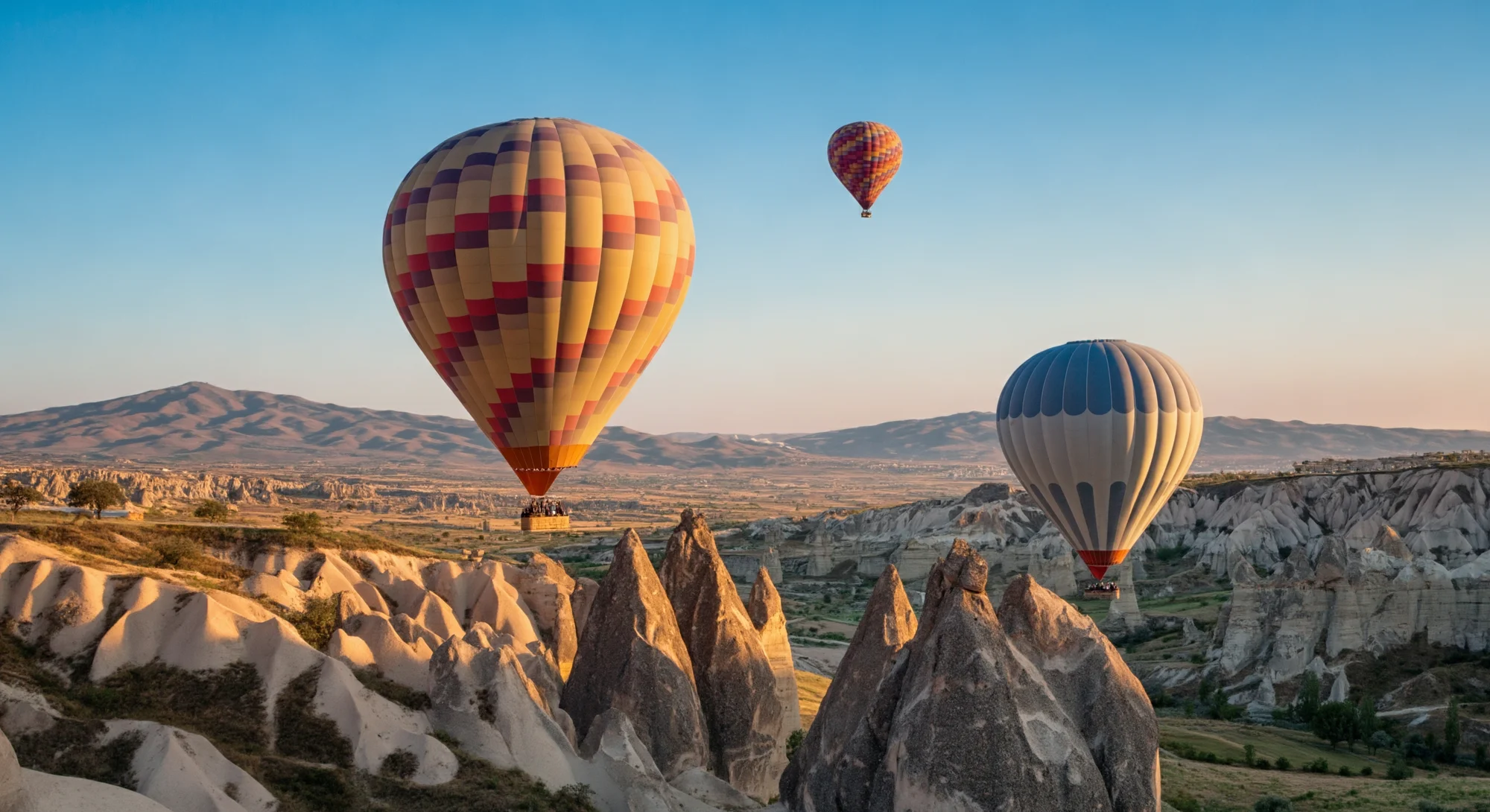 Three hot air balloons float in the sky above a rugged landscape of rock formations. The balloons are colorful and have a basket hanging below them. The sun is shining and the sky is blue.