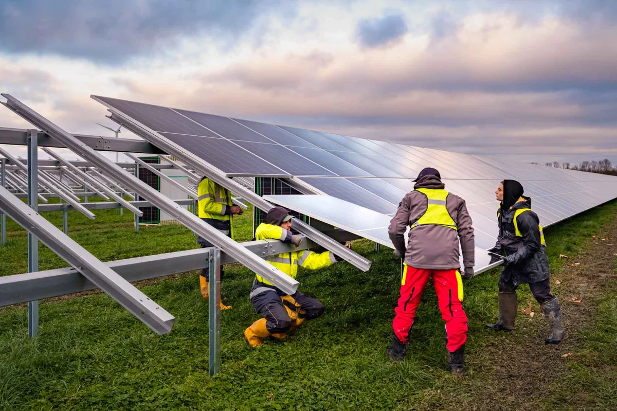 Four people wearing high-visibility clothing are installing large solar panels.