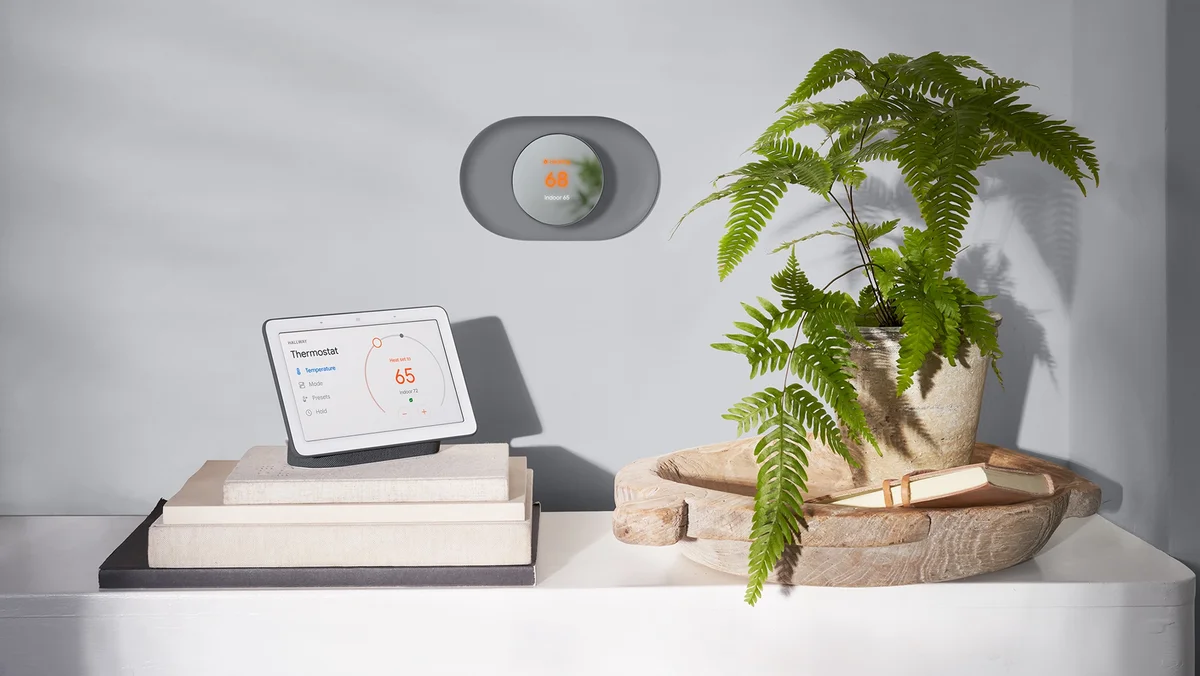 A gray Nest Thermostat hangs above a white table. A green plant and a Nest Hub sit on the table.