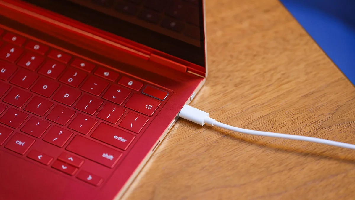 An open, red Chromebook is positioned on the left side of the frame of a photo, revealing about half of the right side of its keyboard. A white cord stretches across from the right side of the image, plugged into the Chromebook.