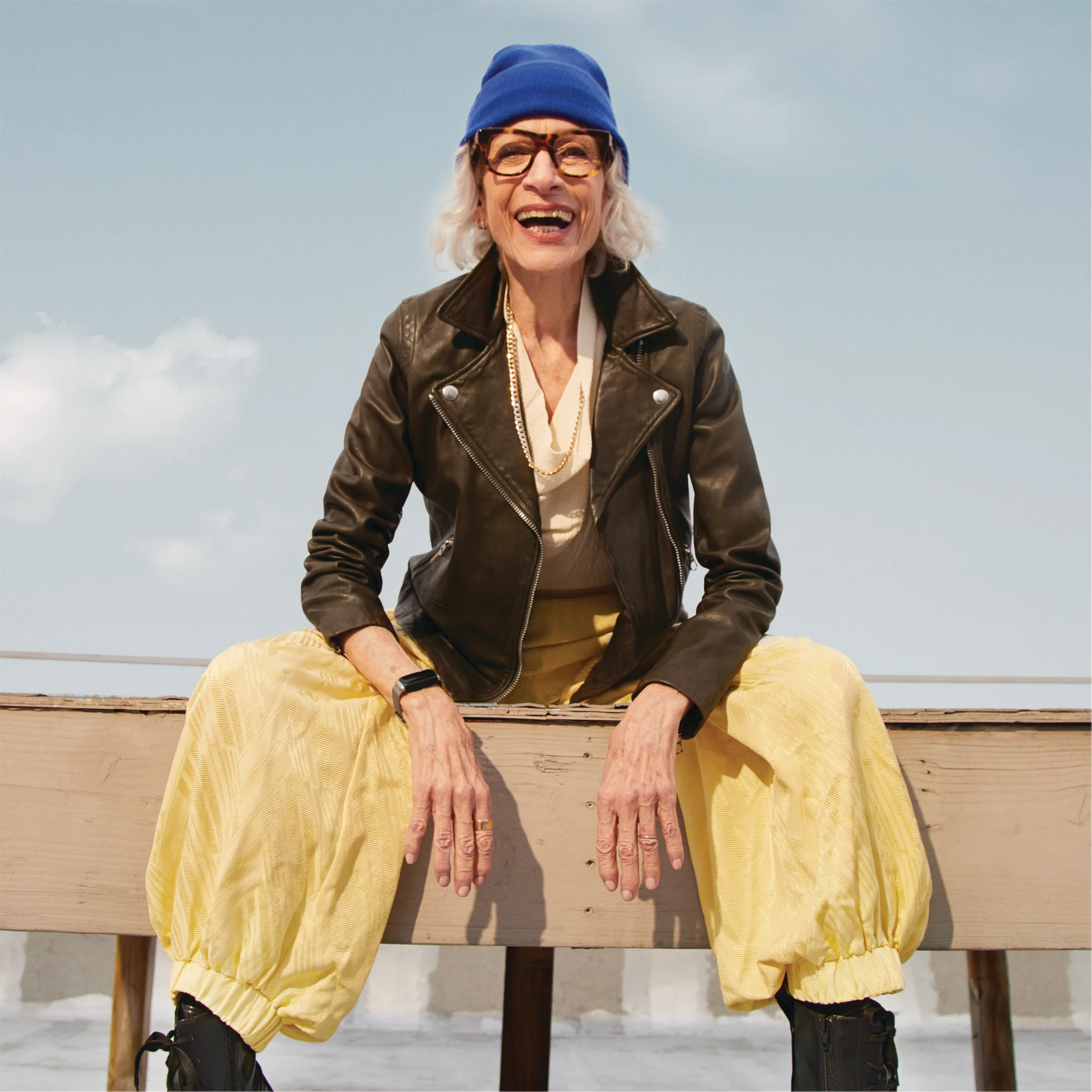 A photograph of an older woman in a leather jacket and a beanie sitting on a bench smiling while wearing Inspire 3.