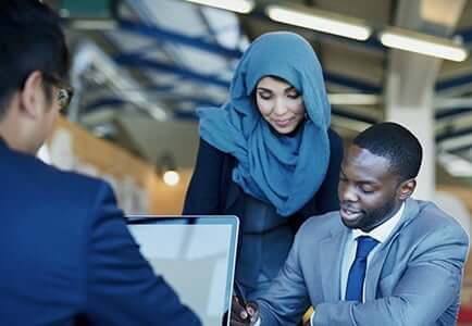 female employee in a hijab looking over the shoulder of a male employee at his desk