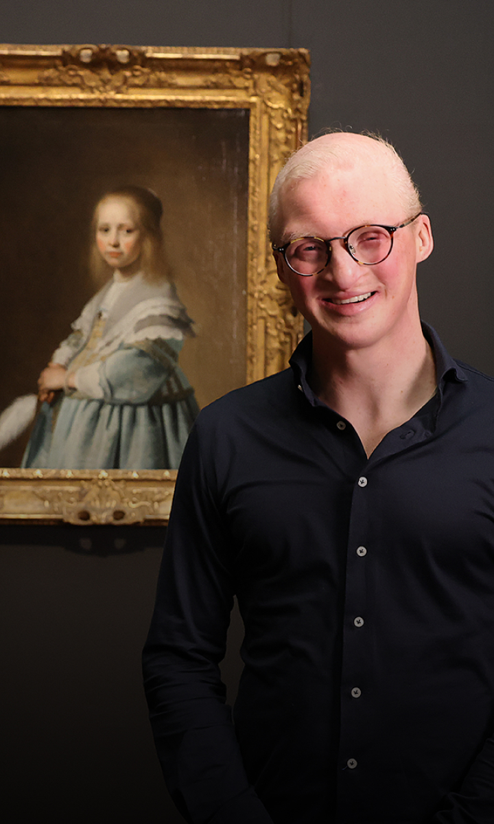 Young man with albinism smiling, standing next to a framed portrait of a young girl in a museum.