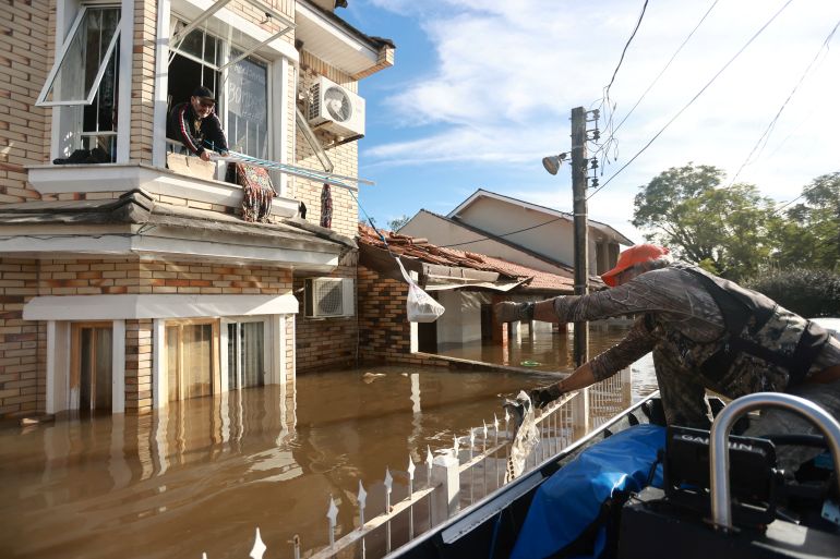 A man reaches across flood waters to a person leaning out of the window of the second story of a house, who appears to be lowering a ladder onto the roof below. There is a brown river of floodwater separating the two people where a road would ordinarily be.