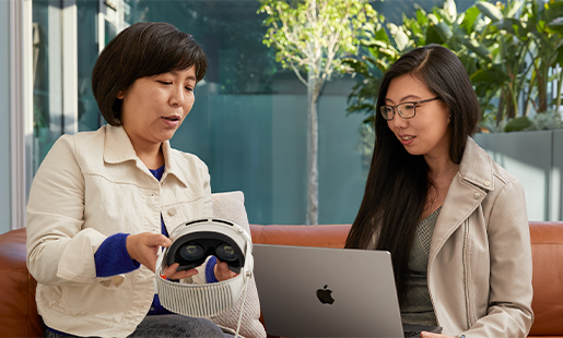 Jessica sitter med en Apple-kollega som visar henne sin Apple Vision Pro. Framför sig har hon en MacBook och i bakgrunden står en dörr ut mot terrassen öppen.