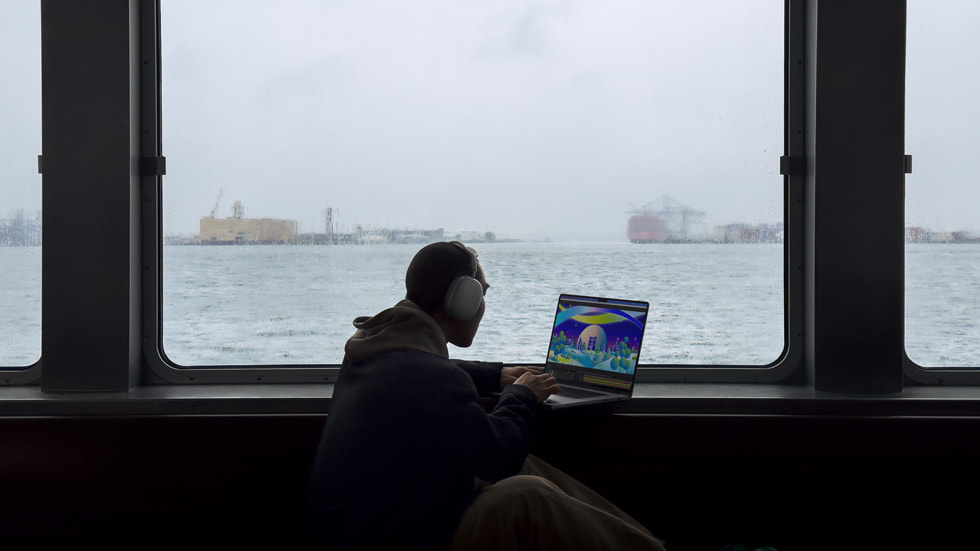 Pessoa com fones de ouvido usa o novo MacBook Pro ao lado de uma janela com vista para um porto.