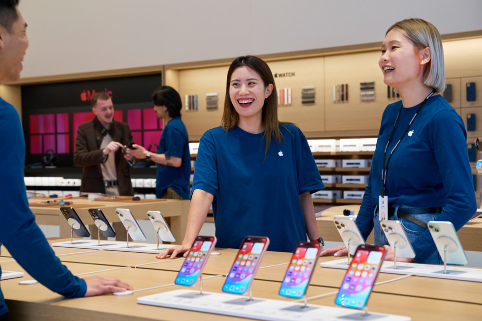 최신 iPhone 제품군이 진열된 테이블 주변에 자리한 세 명의 Apple Store 팀원 모습.