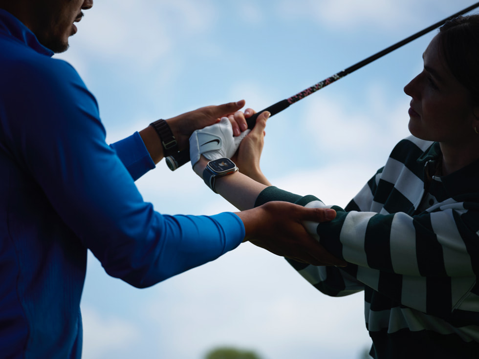Hình ảnh huấn luyện viên golf đang giúp golf thủ đeo đồng hồ Apple Watch thực hiện kỹ thuật vung gậy đánh golf. 