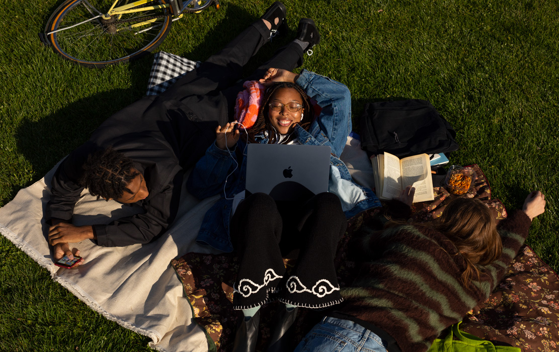 Three university students lay on a blanket in the park. One student has an iPhone. One student has a MacBook Air and Apple headphones. One student has a paperback book.