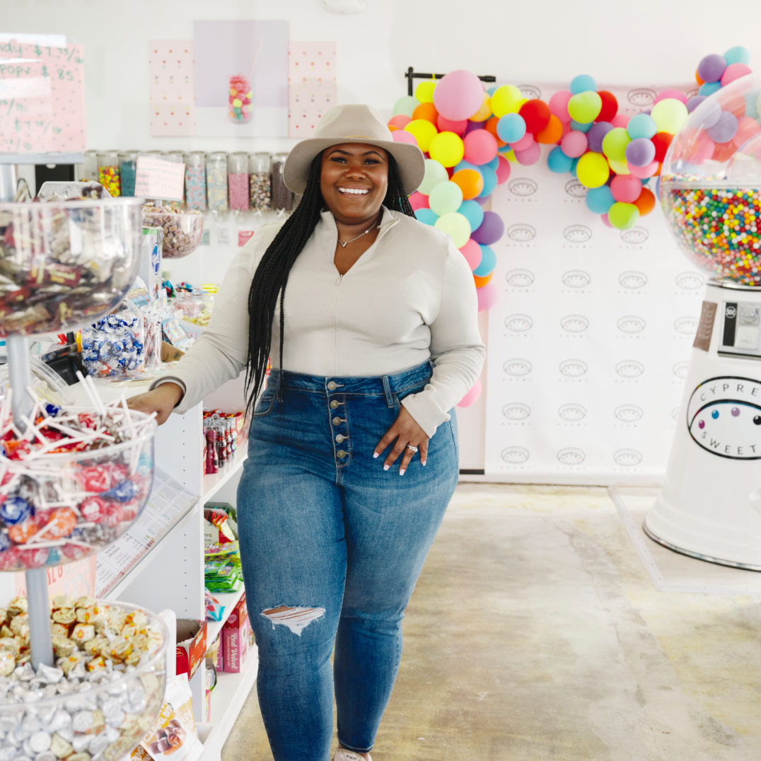 The business owner of Cypress Sweets standing and posing for a photo in her candy store, wearing casual clothes and a hat.