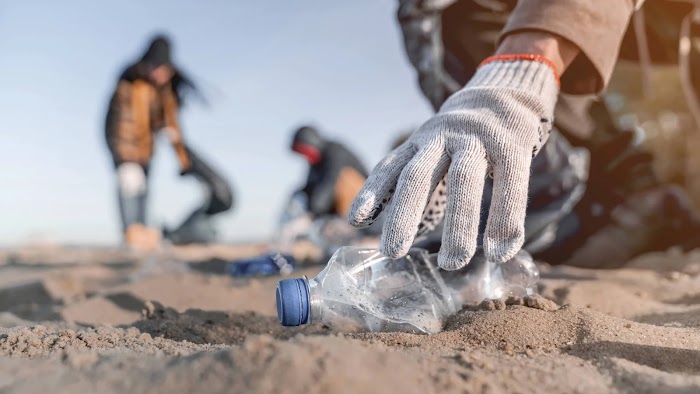 Una mano enguantada se extiende para tomar una botella de plástico en una playa.