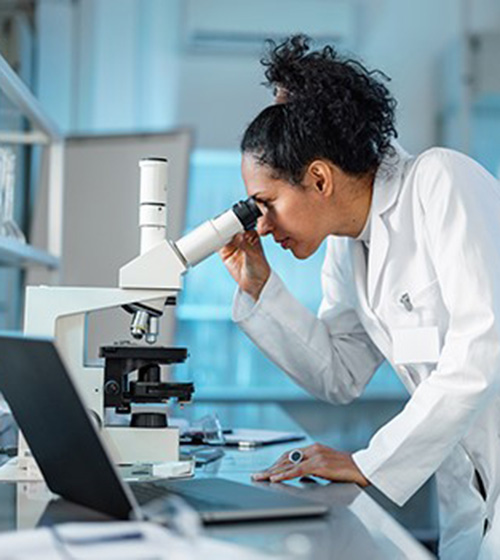 woman in labcoat using a microscope with a laptop nearby