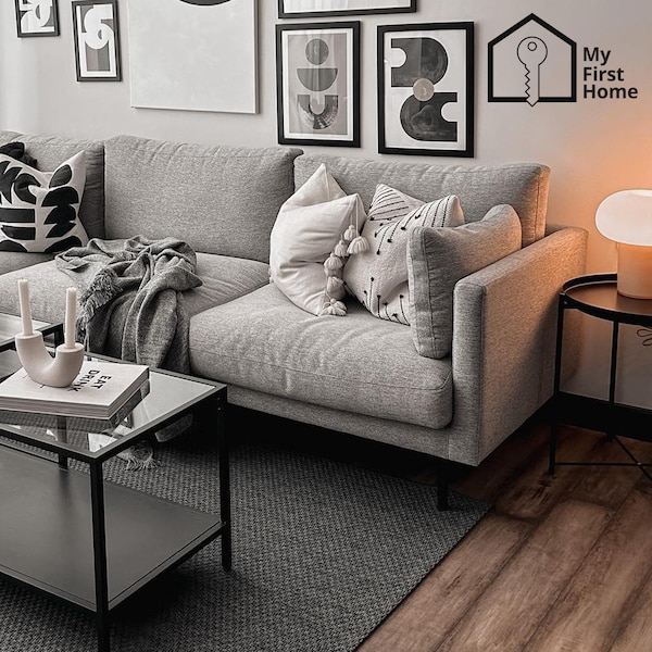 A grey SÖDERHAMN sofa is placed on top of grey rug in the living room, in front of a rectangular VITTJSÖ coffee table. Pictures in black frames are hung on the wall behind the sofa. Beside the sofa, a DEJSA table lamp placed on a black GLADOM table i