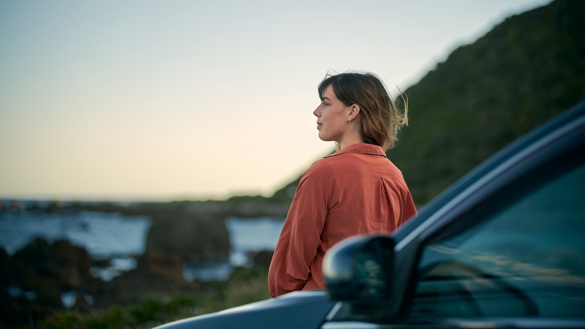 A woman stands beside her car and looks out at the water. She is calm