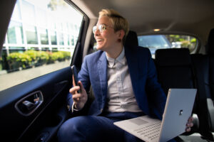 a person with short haircut and a suit sitting in a car looking at a laptop