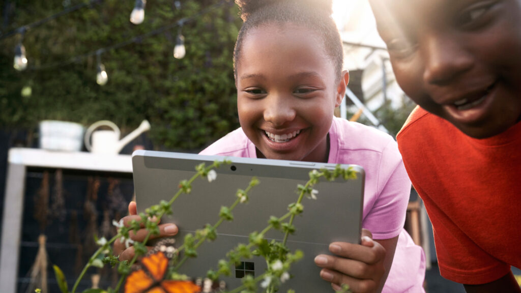 Two children engaged, looking at a tablet while outdoors in a grassy area.